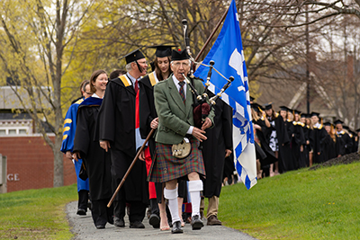 A bagpiper leads a procession of robed faculty members and students down a path towards Convocation Hall