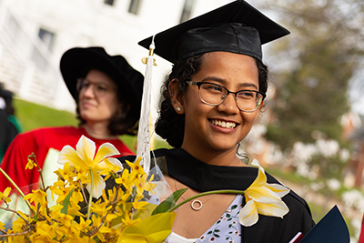 A female graduate smiles while holding her diploma and a bouquet.